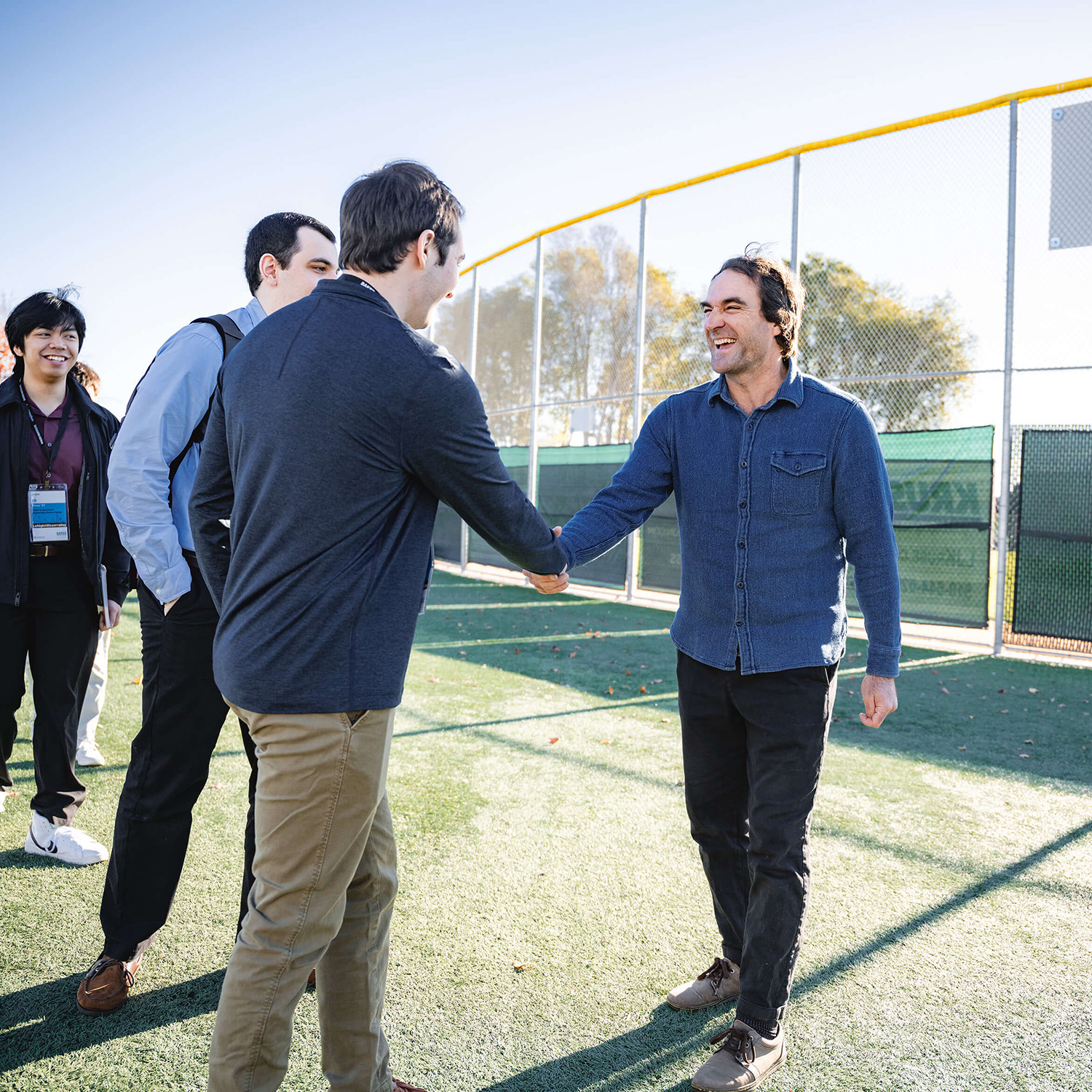 Two men eagerly shake hands while outside on a turf field with big smiles.