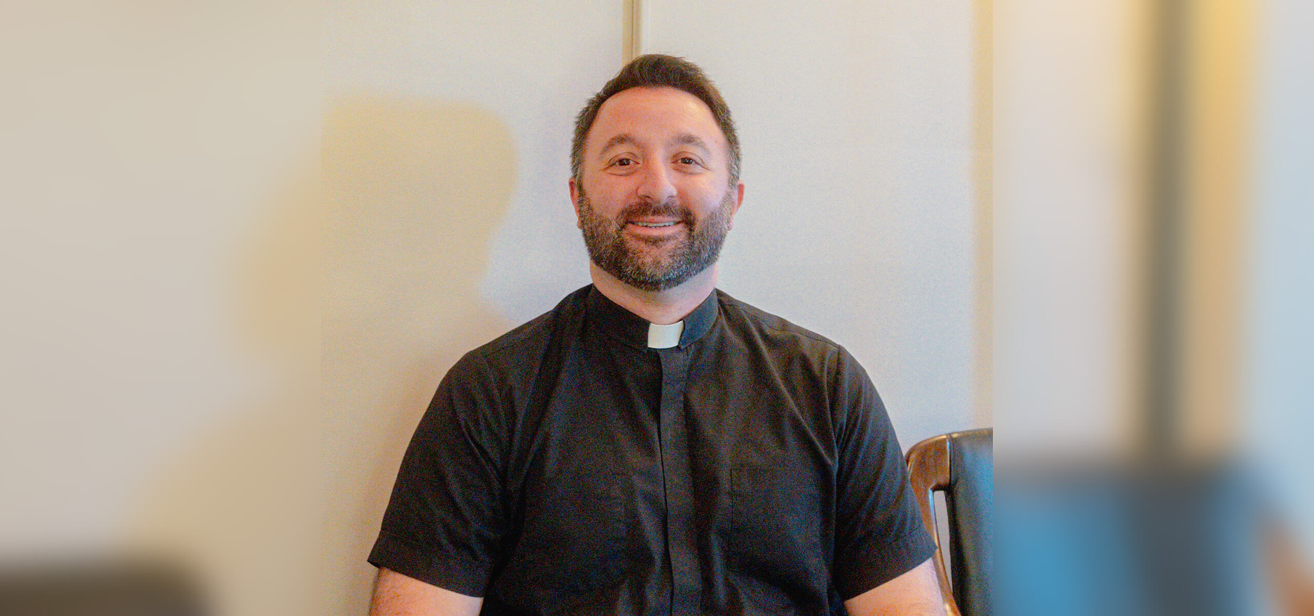 A smiling man with dark hair and a short beard wears a priest’s collar and black short-sleeved shirt.