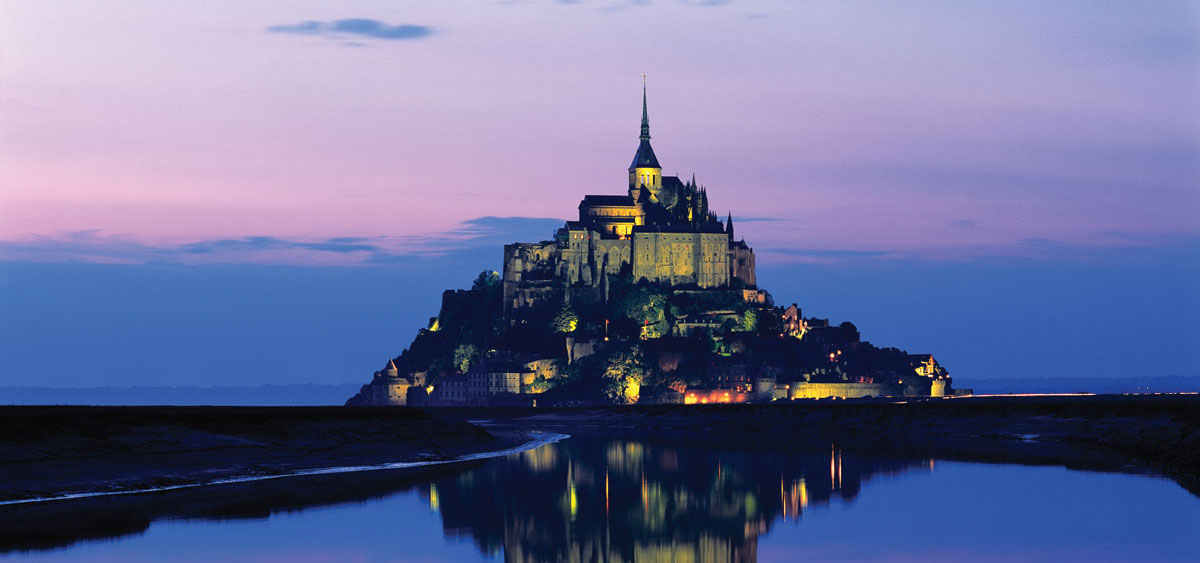 Mont Saint-Michel at night, overlooking mirror-like water.