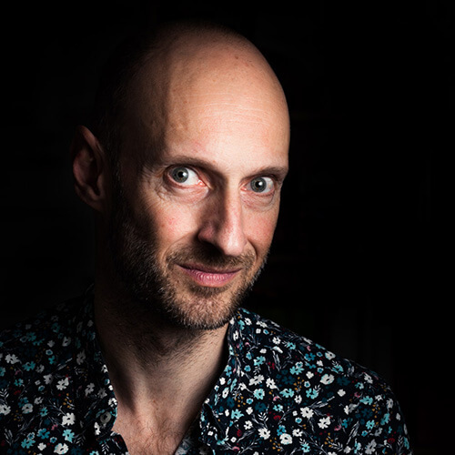 In a dark studio, a headshot of Franck Vogel bearing a closed mouth smile and wearing a black shirt with blue print is heavily shadowed.