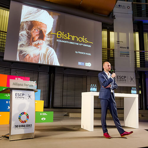 Standing on a stage with a large monitor behind him, Franck Voge speaks in a navy suit and holding a microphone.