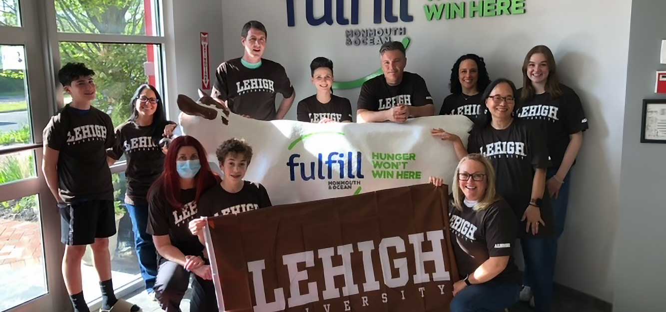 Alumni and friends wearing brown Lehigh shirts huddle together. A few alumni are holding a Lehigh flag and a banner reading "Fulfill, Hunger won't stop here."