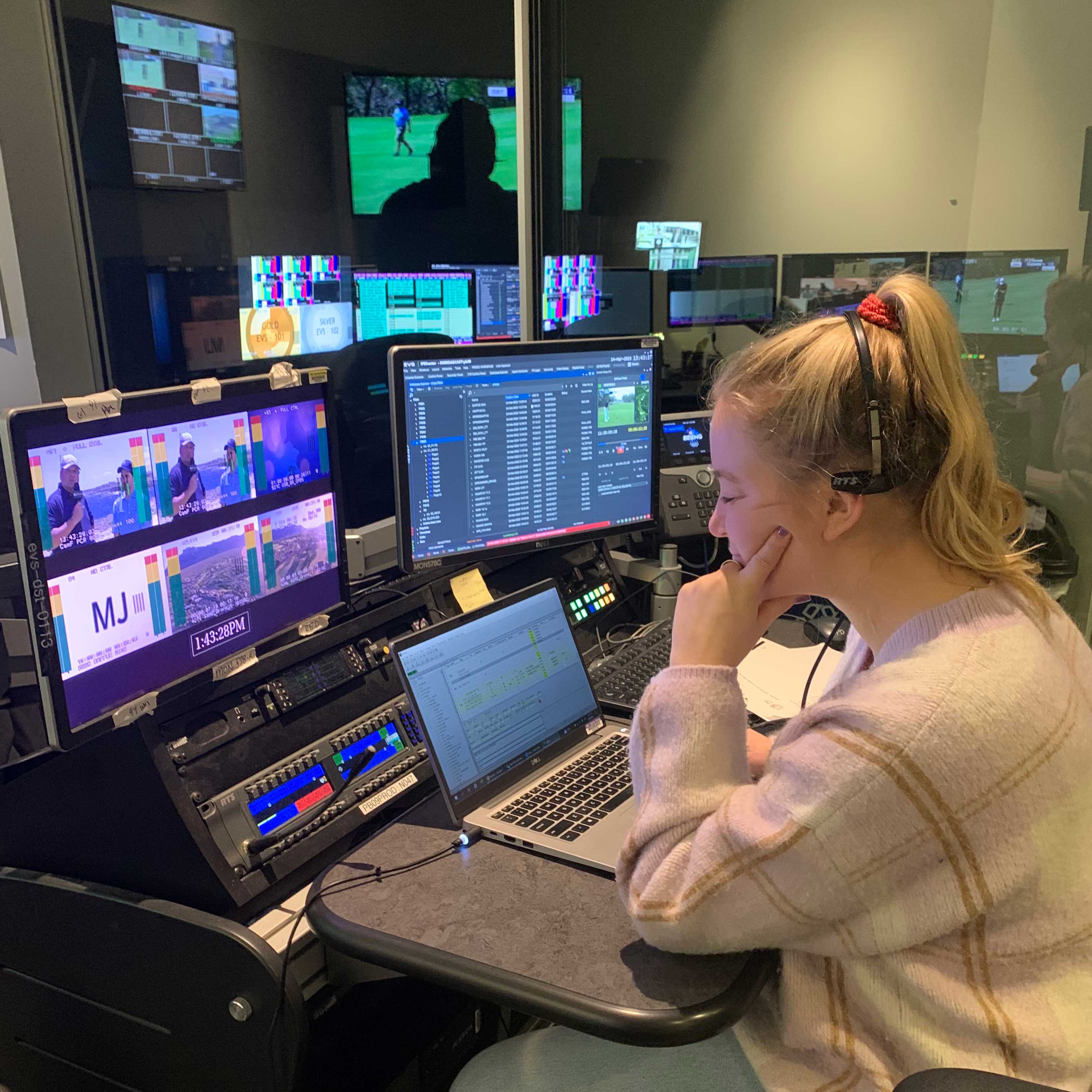 Madison sits in a production room with several screens in front of her. She watches footage and listens with a headset.