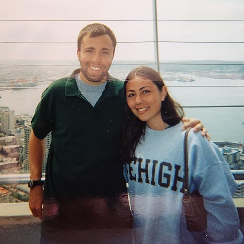 Leili and Brian McMurrough from their Lehigh days. They stand atop a tall building with water in and background.