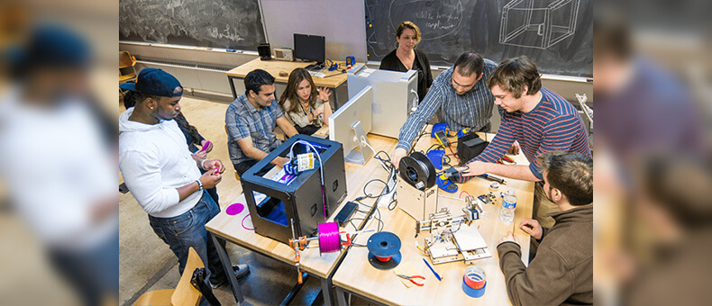 A group of students and a professor gathered around a 3D printer, working together and sharing ideas on their printing project