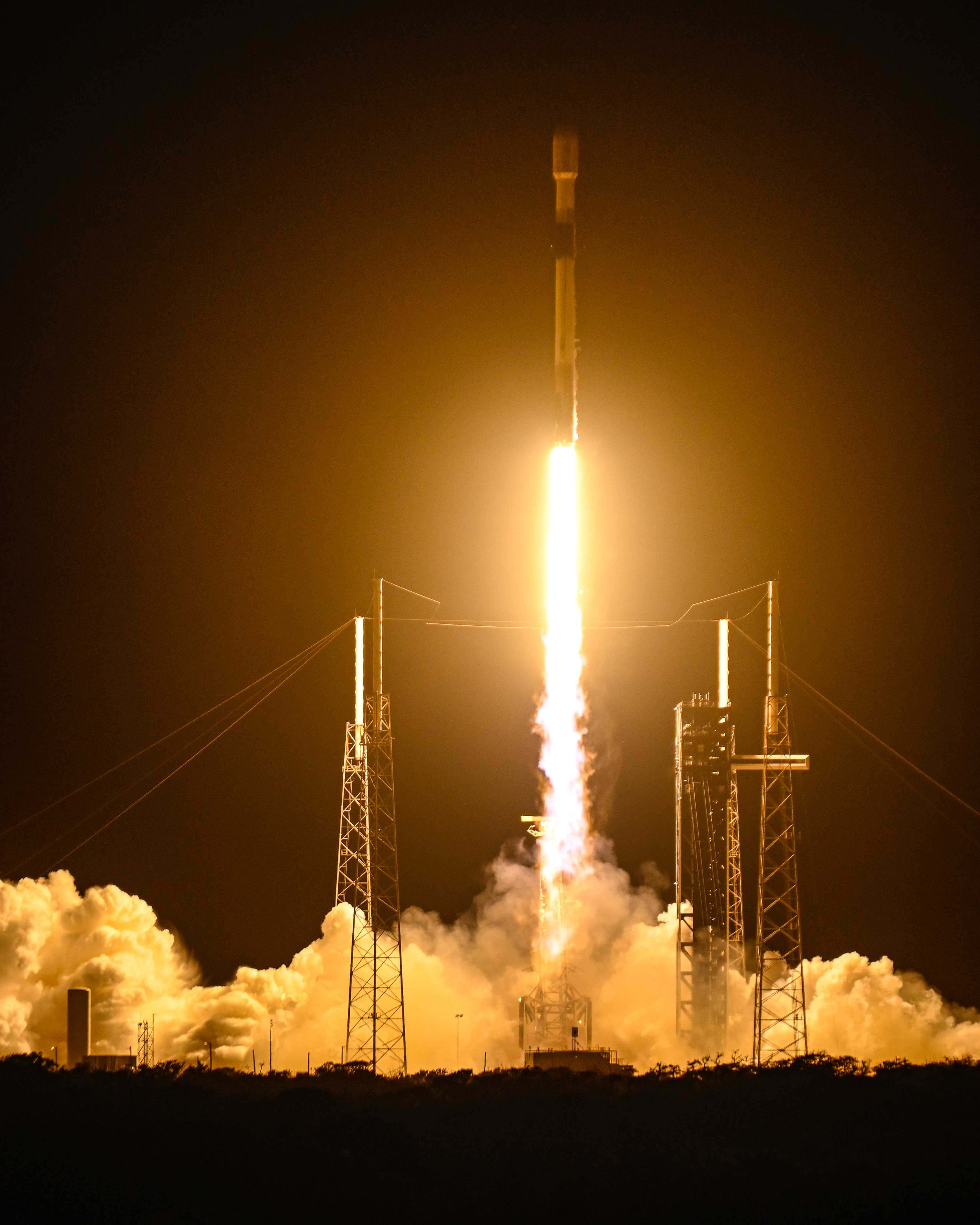 GPS III Sv10 mid-launch. Bright orange and yellow exhaust gas burns as it takes off the launch pad. 