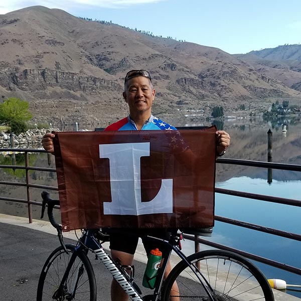 Gary Pan holds up a Lehigh flag while he leans on his bike with a mountain behind him