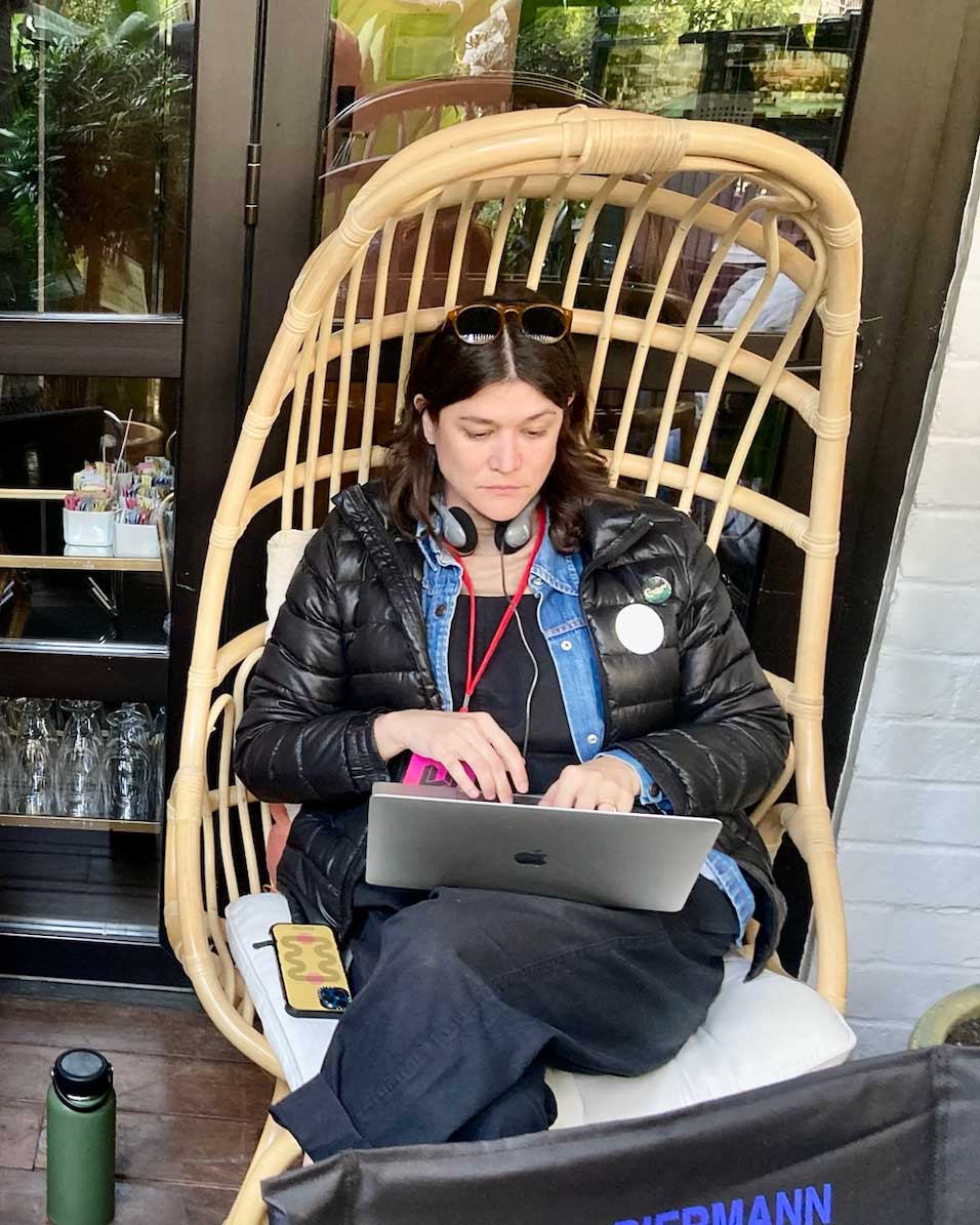Genevieve sits in a wooden, egg-shaped chair with a laptop balanced on her legs. She types; headphones are around her neck.