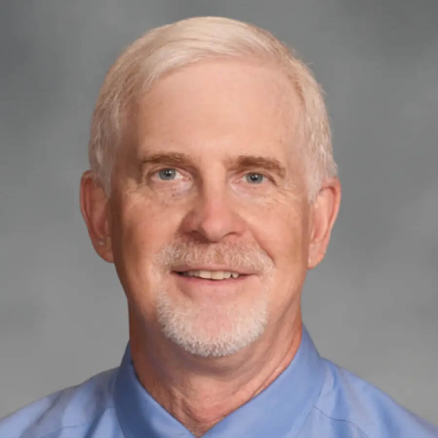 Headshot of a male with white hair and goatee wearing a blue buttoned up dress shirt, posed against a light gray background.