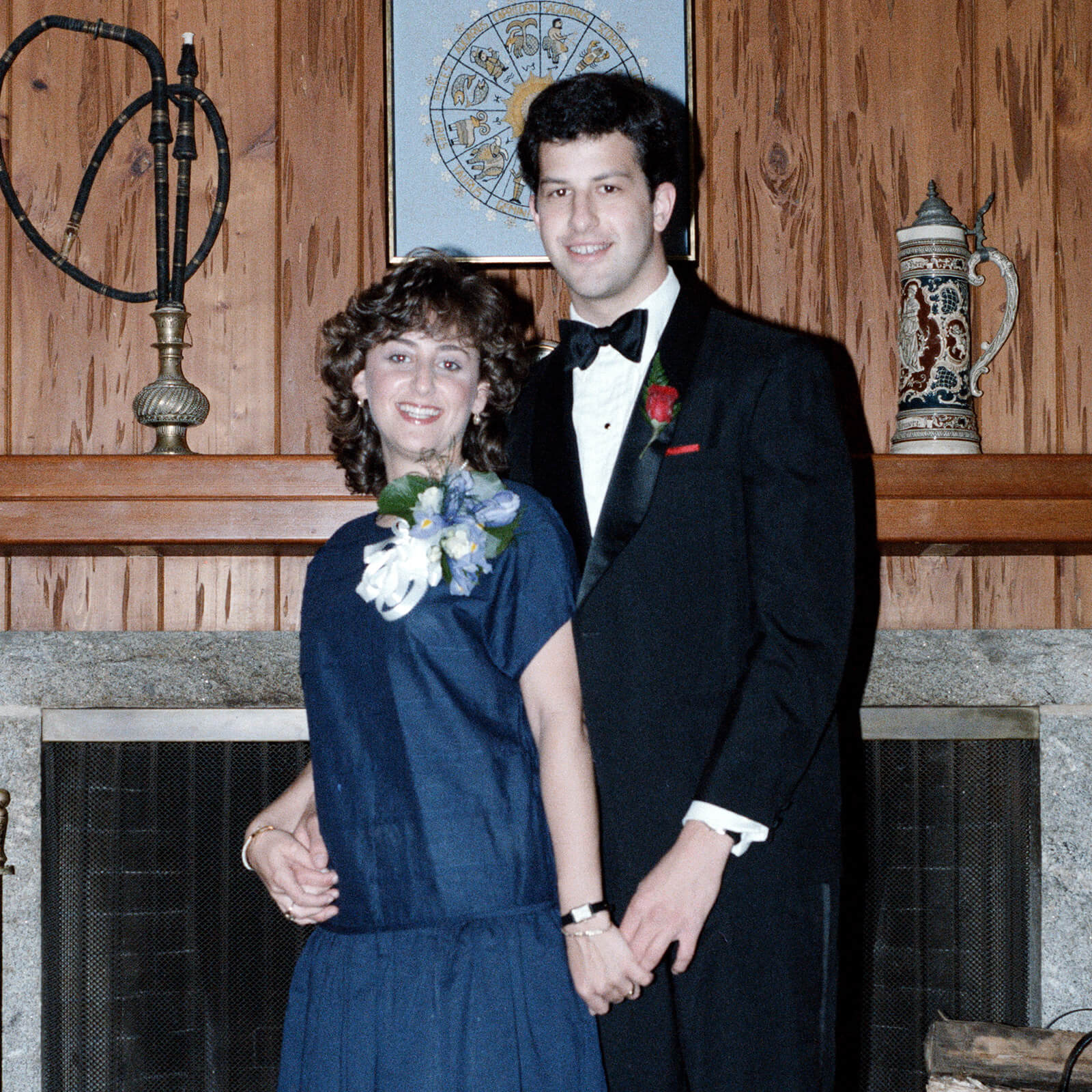 Faith Glazier and Robert Weisstuch  pose in formal wear in front of a fireplace with dark wood panelling while each wearing a corsage and boutineer.