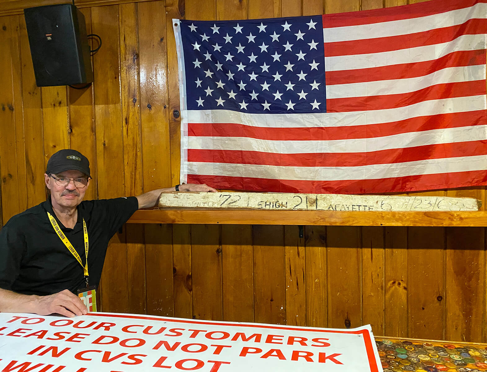 At a wood panelled wall with a large American flag hanging, a man poses with a plank of wood signed in marker with “’72 Lehigh 21 Lafayette 6 11/23/68.”