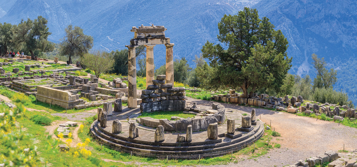 Ruins on a Grecian cliffside surrounded by green grass and other foliage.