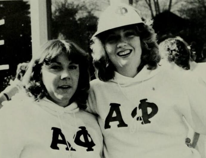 In a black and white photo, two women stand shoulder-to-shoulder wearing white hooded sweatshirts with greek letters on the front.