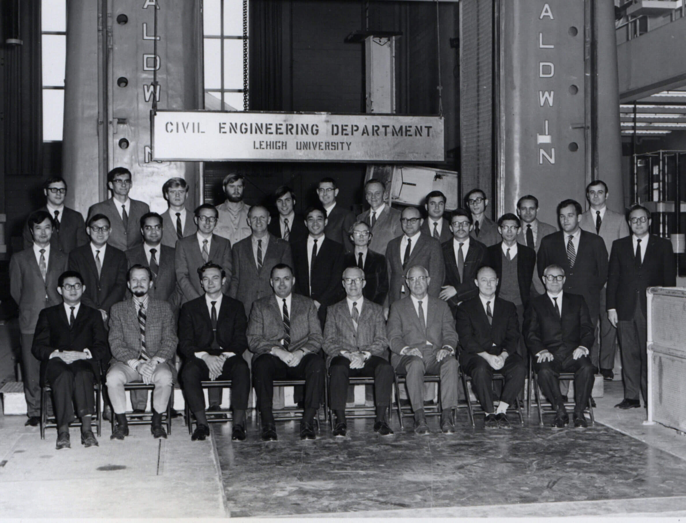 Three rows of men in suit jackets and neck ties pose for a group photo in front of an industrial sized machine, the front row seated in chairs with others standing behind them.