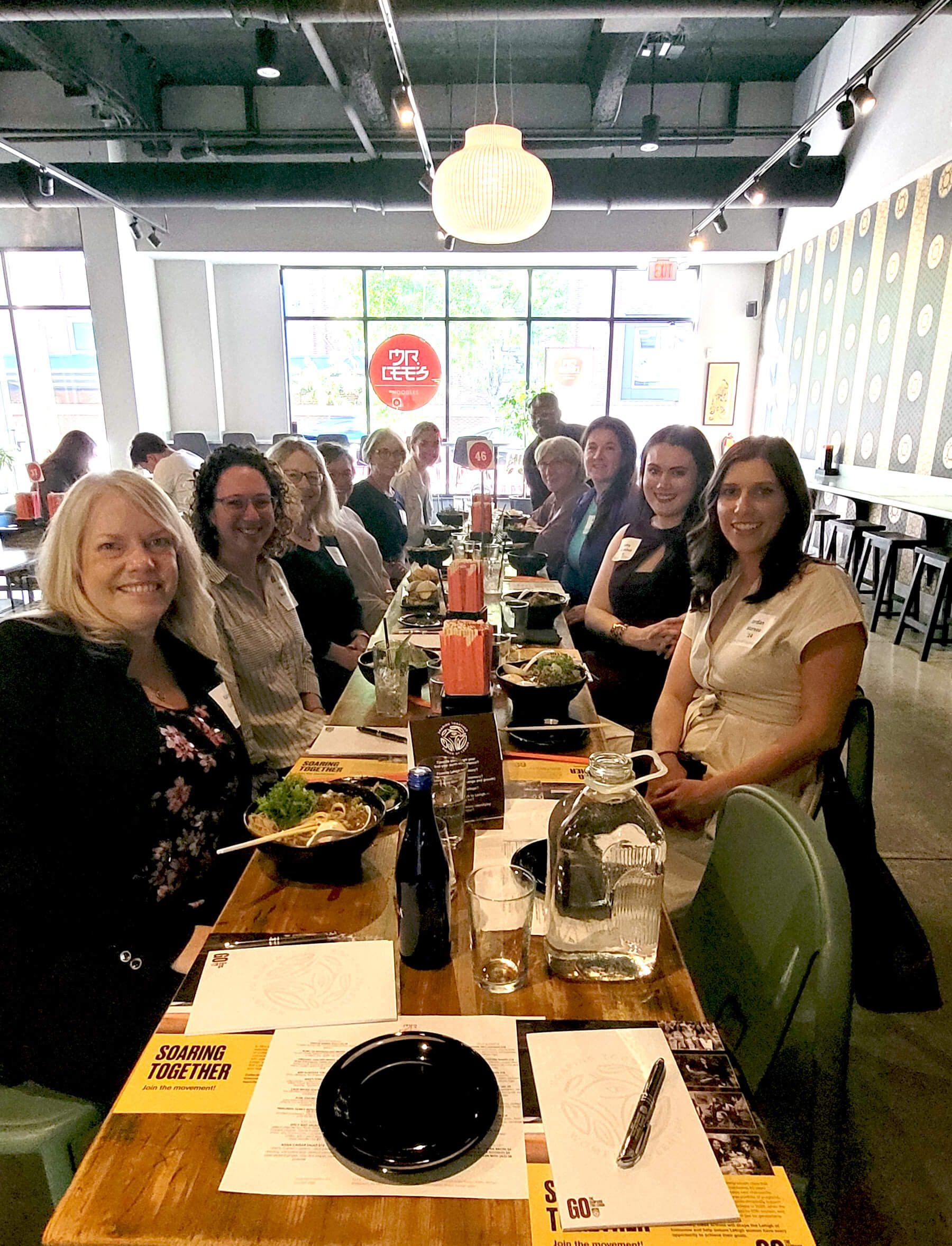 A table of women smile and pose for a photo with a sign on the window of the restaurant in the back reads "Mr. Lees'"