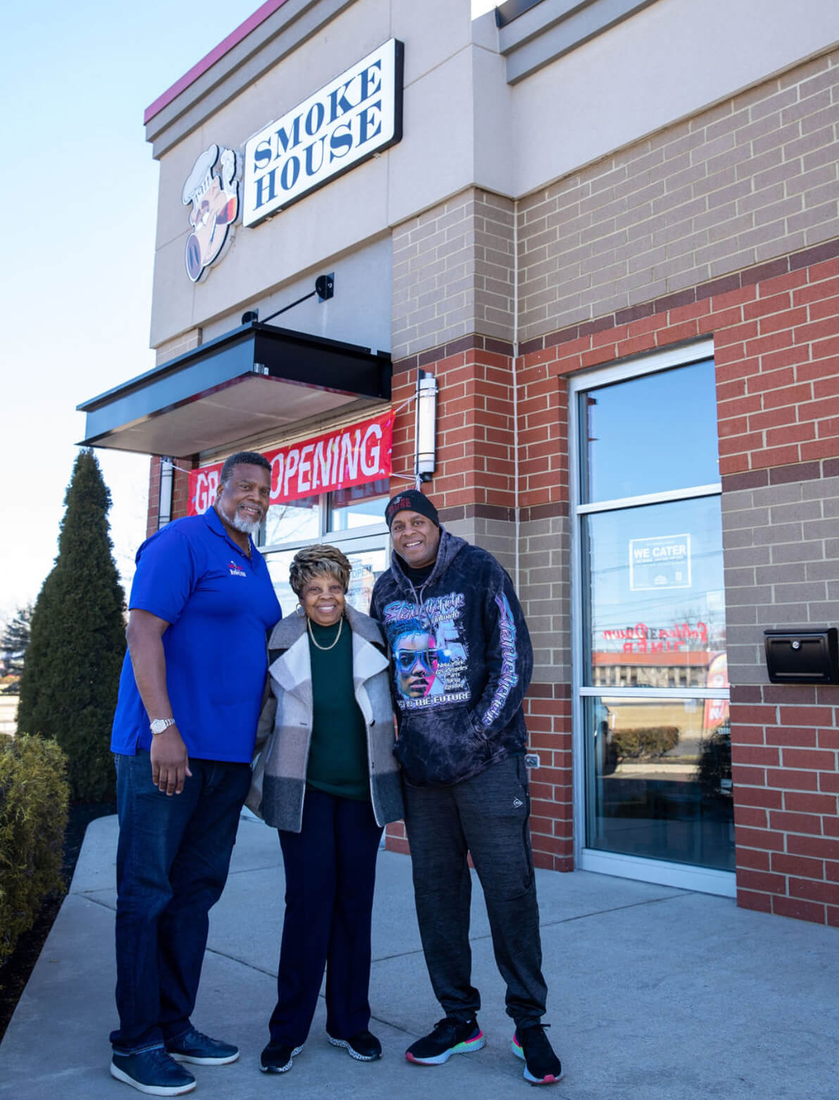 Two tall men stand in front of a grand opening sign at their restaurant with their mother standing between them.