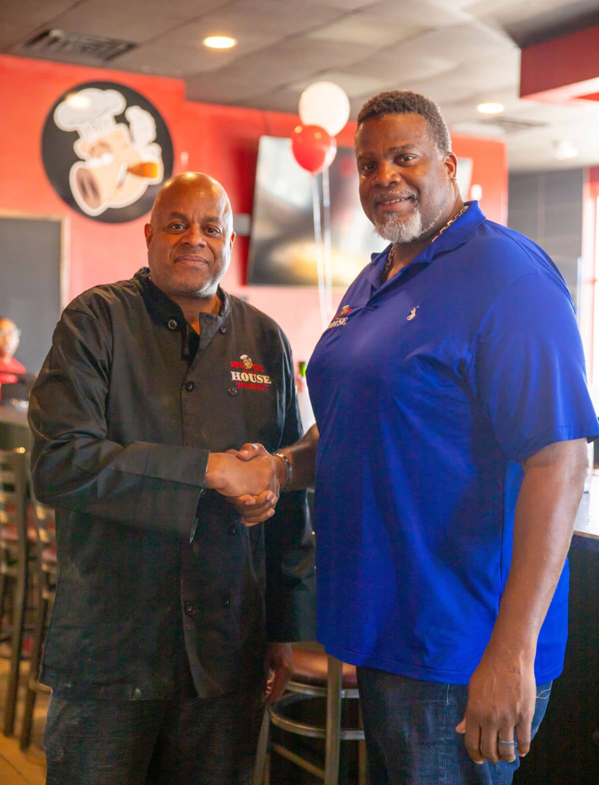 Two brothers stand inside their restaurant shaking hands and smiling at the camera.