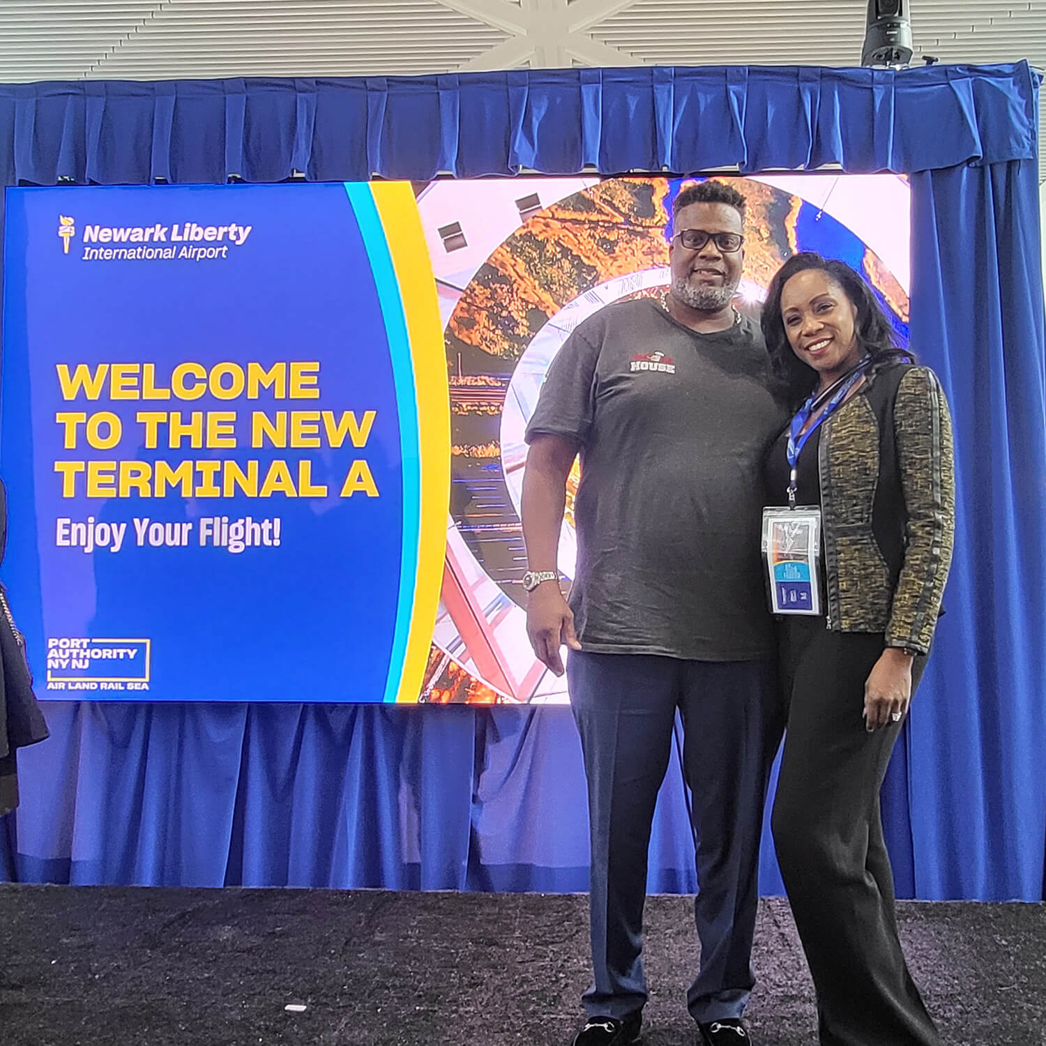 Man and his wife pose at the airport, standing in front of a blue curtain with a sign that says "Welcome to the new terminal A".