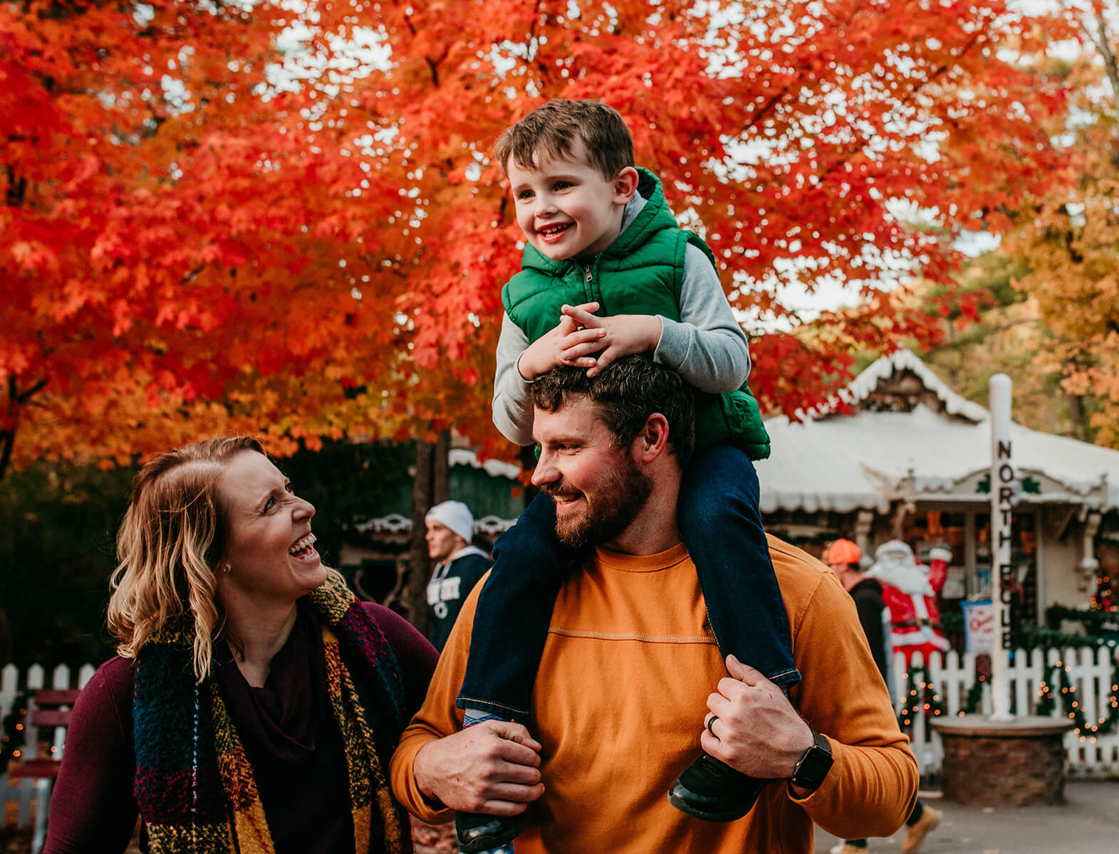 A mother looks up at her son sitting happily on his father’s shoulders with bright orange foliage in the background.