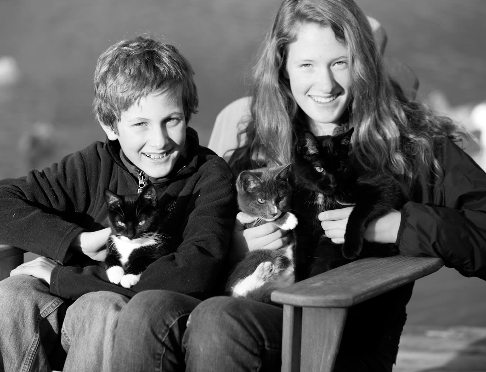 Brother and sister smile at the camera in a black and white photo, holding three, tiny black and white kittens.