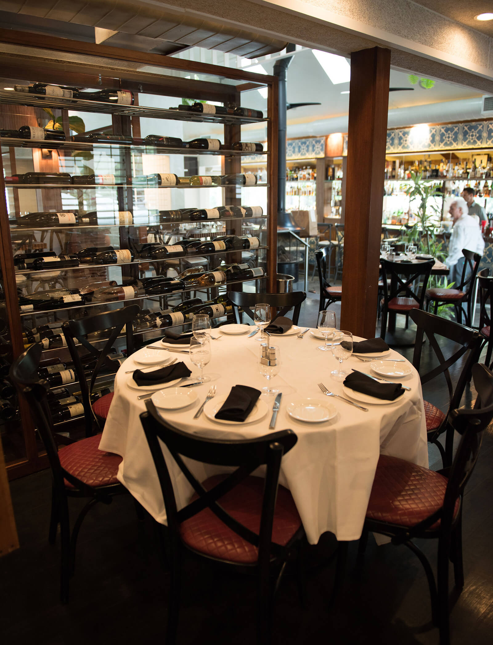 A well set round table with a white table cloth with a floor-to-ceiling wine rack beside it and a bar in the background.