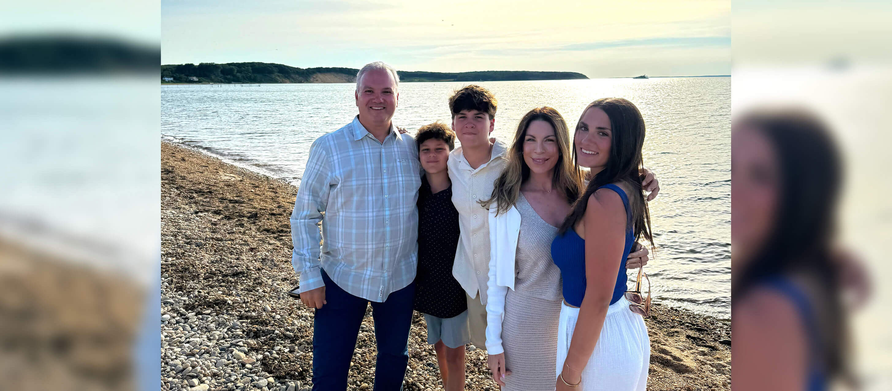 Family of five stands huddled for a selfie on a beach with the water, blue skies, and clouds behind them.