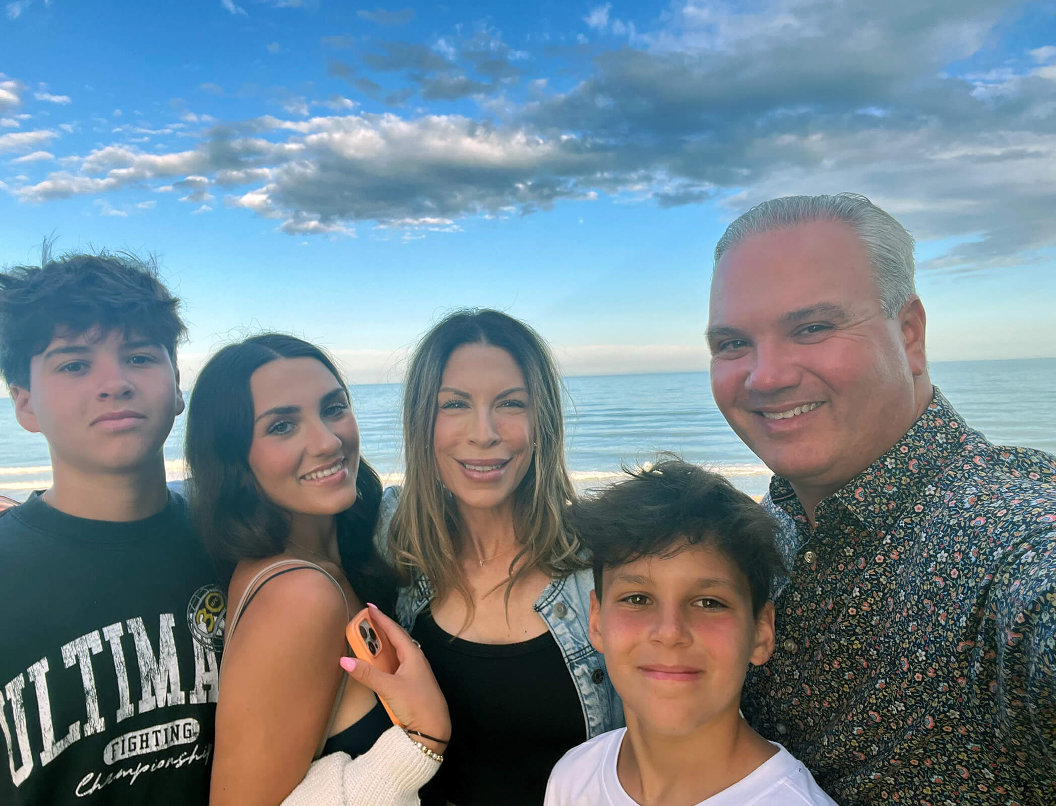 Family of five stands huddled for a selfie on a beach with the water, blue skies, and clouds behind them.