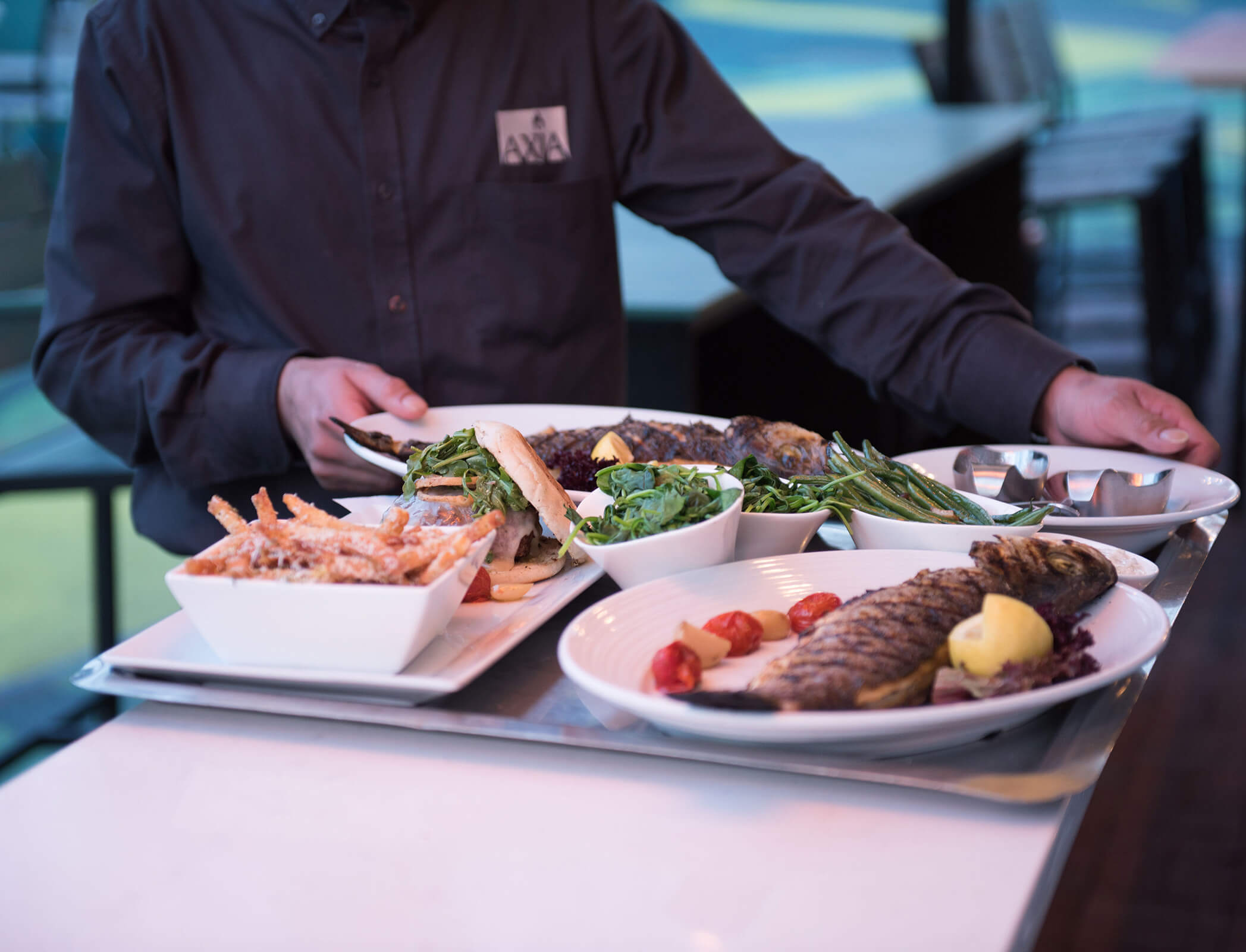 A server wearing a black shirt with an Axia logo places plates of Mediterranean food on a tray.