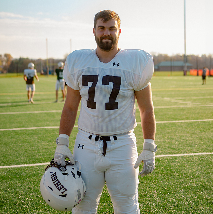 Football player #77 poses on the field, facing the camera holding his helmet at his side.