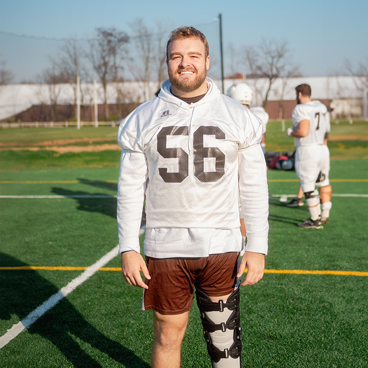Football player #56 poses on the field, facing the camera and wearing a knee brace.