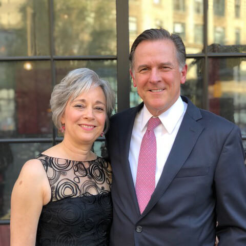 Dressed in semi formal attire, Ann and Weston Hicks pose smiling for a photographer in front of a wooden door with windows.