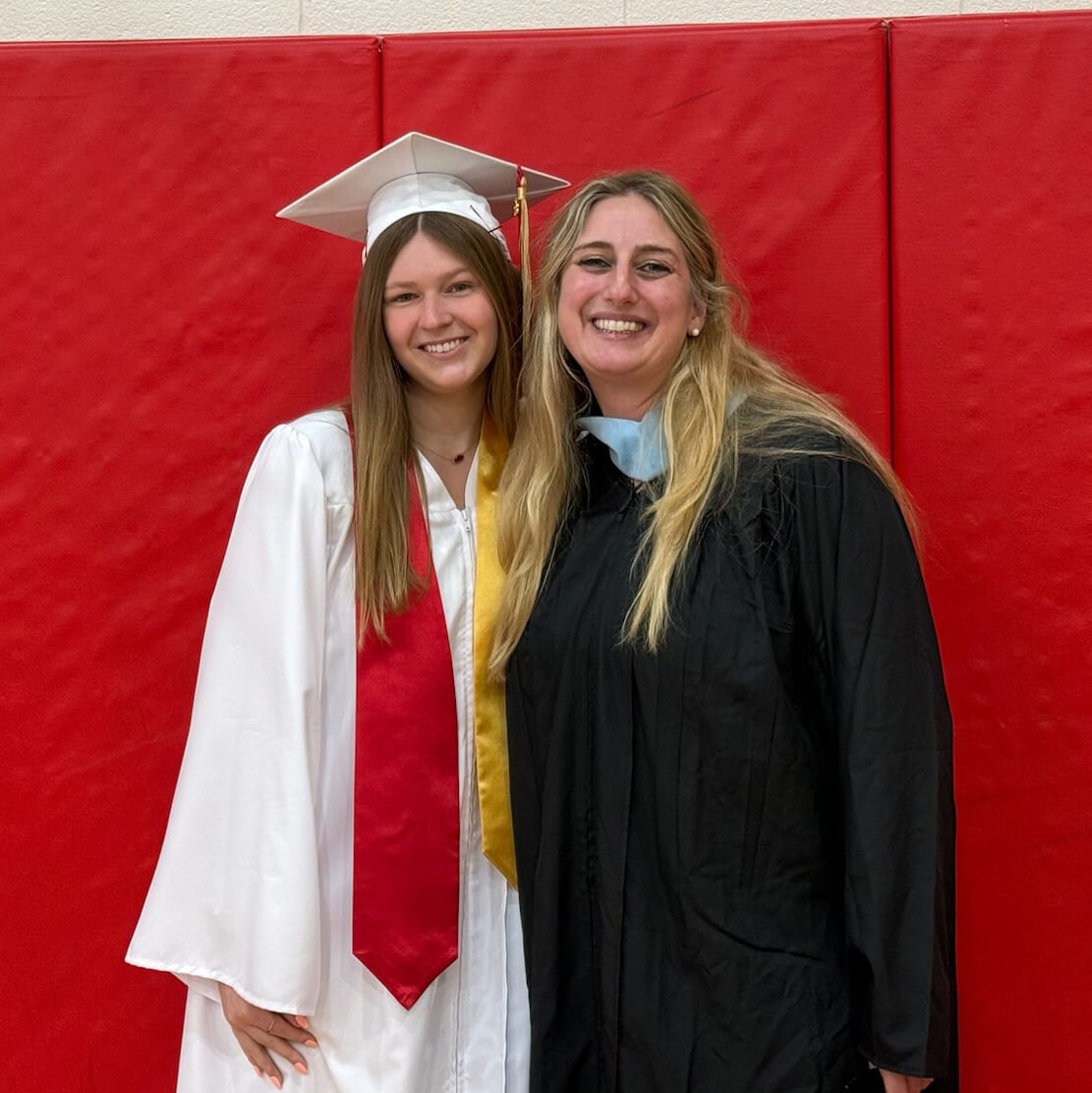 Rochelle Sheridan smiles in a high school gym wearing a black robe posed beside Molly Campbell in a white graduation cap and gown with a red sash.