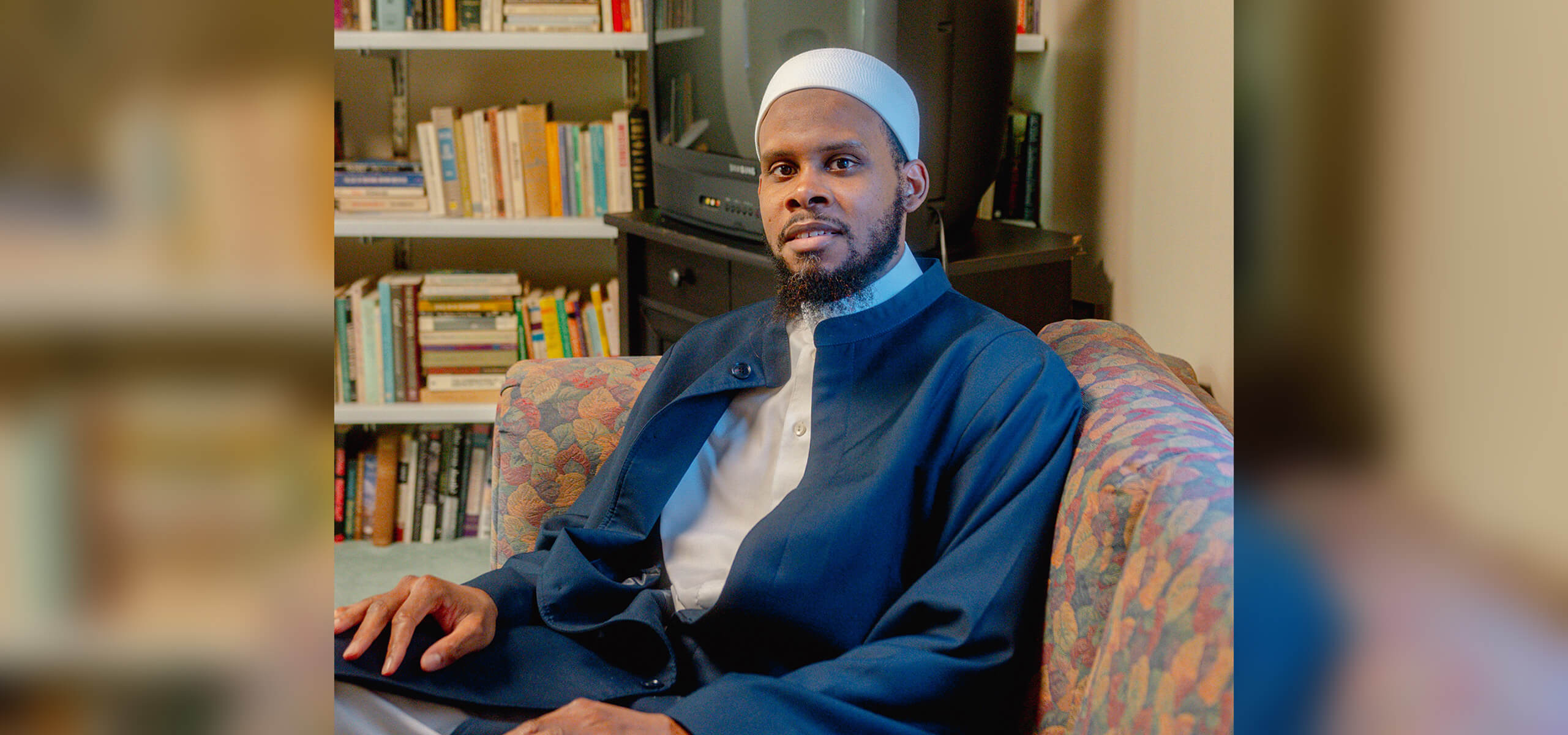 Sitting on a couch with books on a shelf behind him, a man sits smiling and wearing a dark beard and white cap.