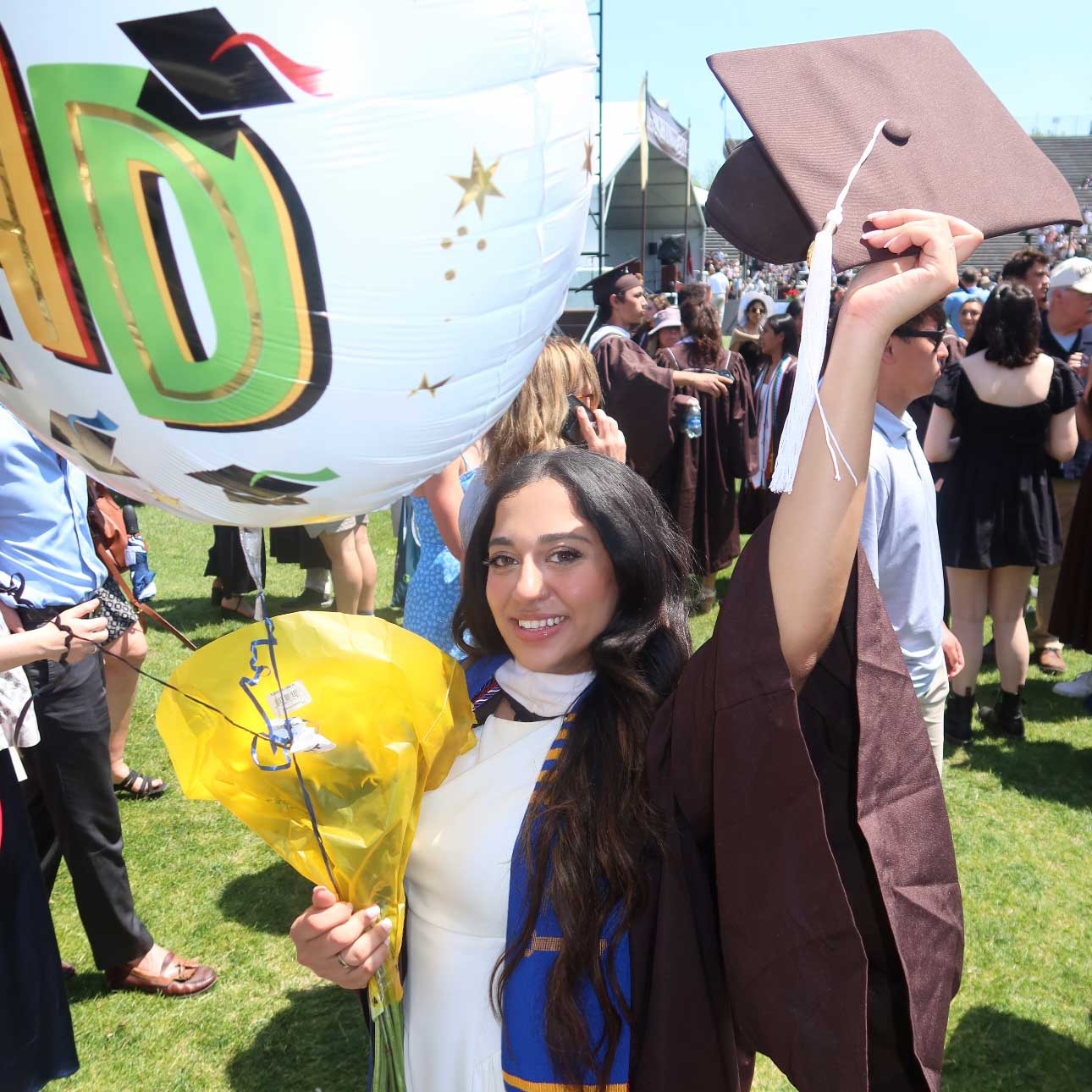 Iqra wears a white dress under her graduation gown. She extends her arm and holds up her cap; her other hand holds balloons.