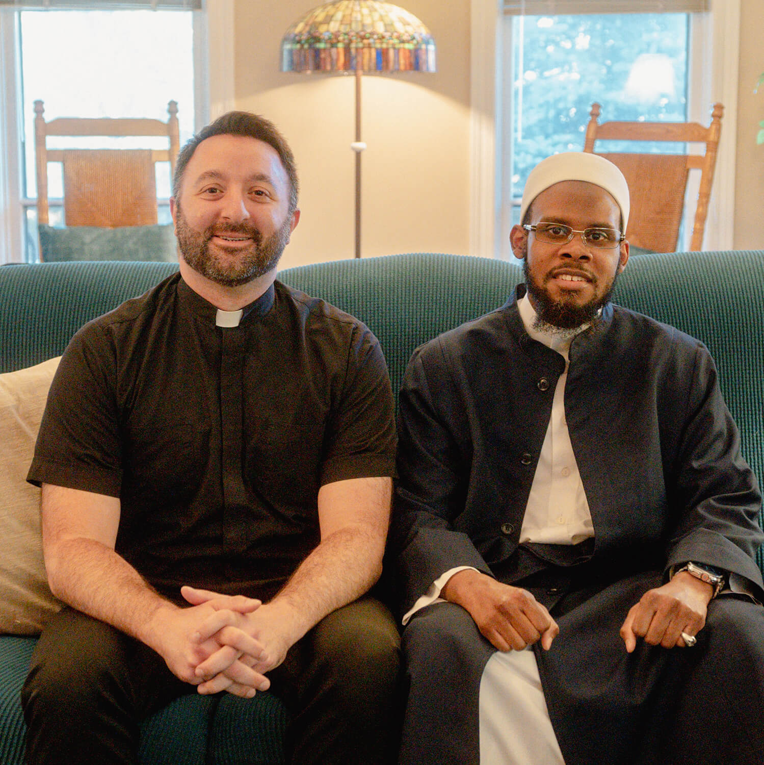 Two men sit on a green couch, both wearing their respective religious garments, on the left a priest and on the right an imam.