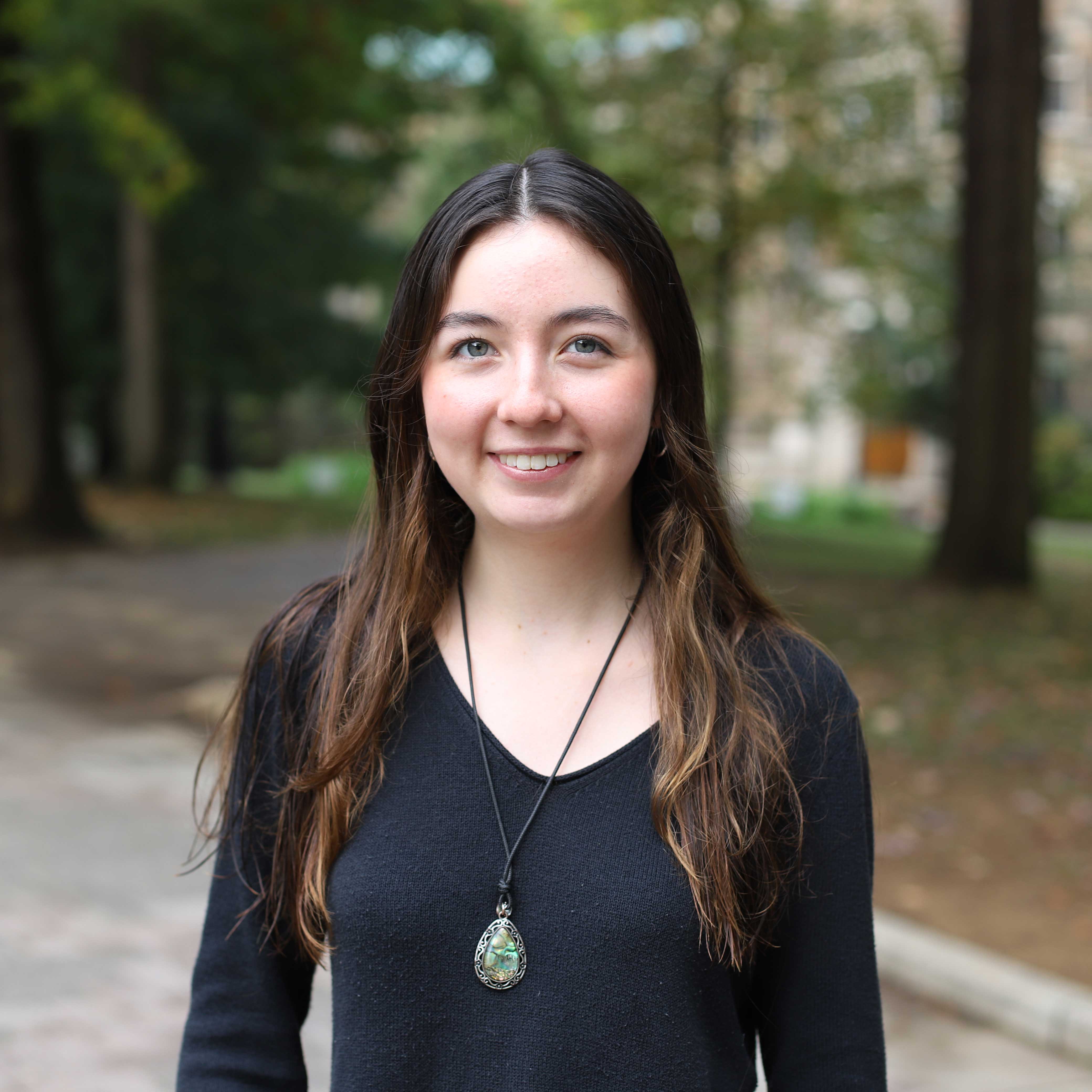 Isabella wears a black long sleeve shirt and green gemstone necklace. Campus is visible behind her.