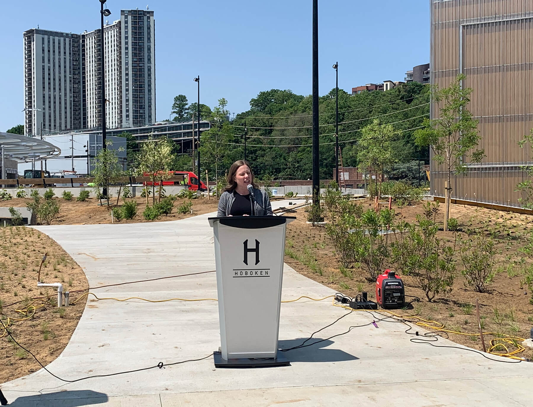 Jennifer Gonzalez stands in a newly planted green space at a podium emblazoned with a large letter H and the name Hoboken.