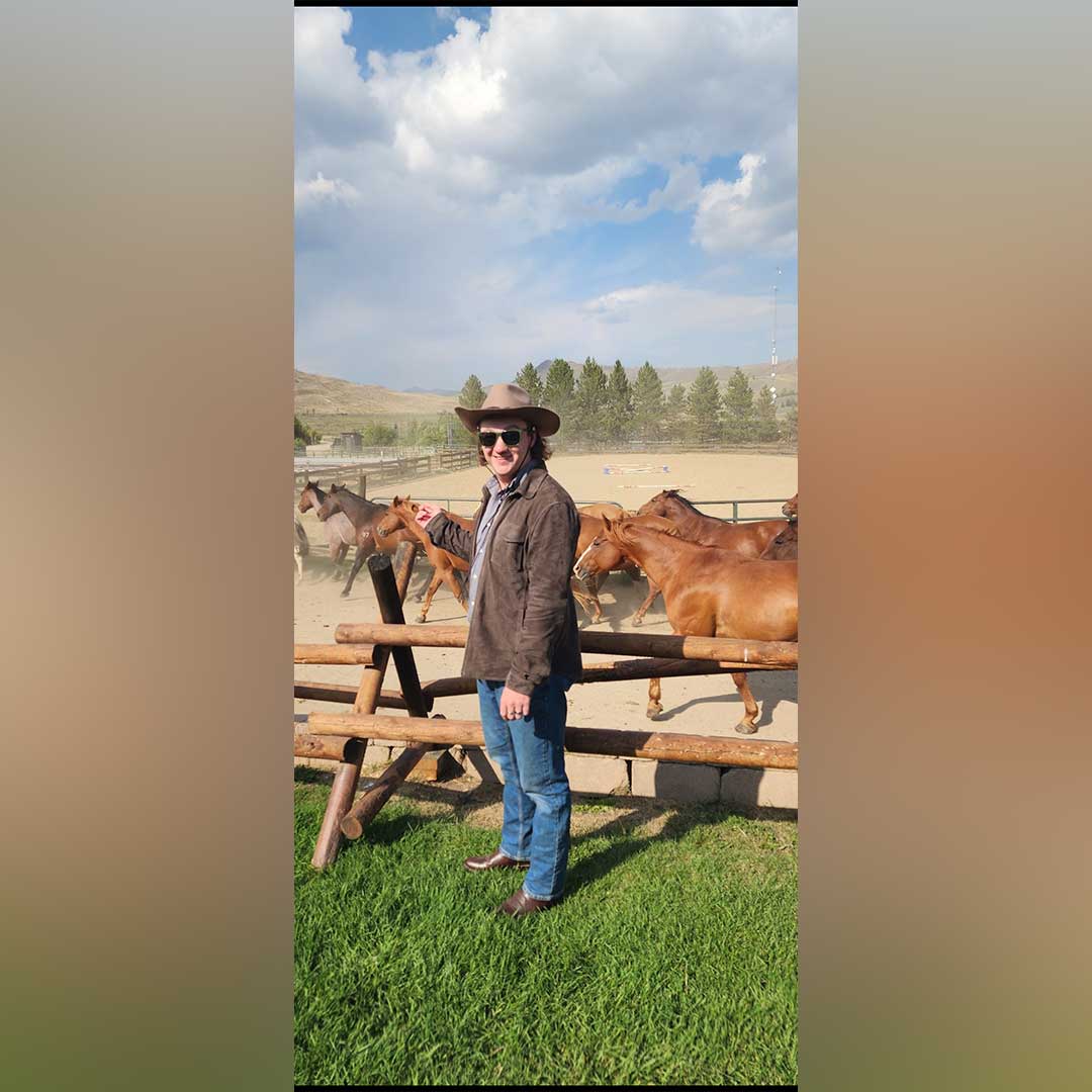 JP stands next to a wide, open area with horses running around. He wears a cowboy hat and a brown barn jacket.
