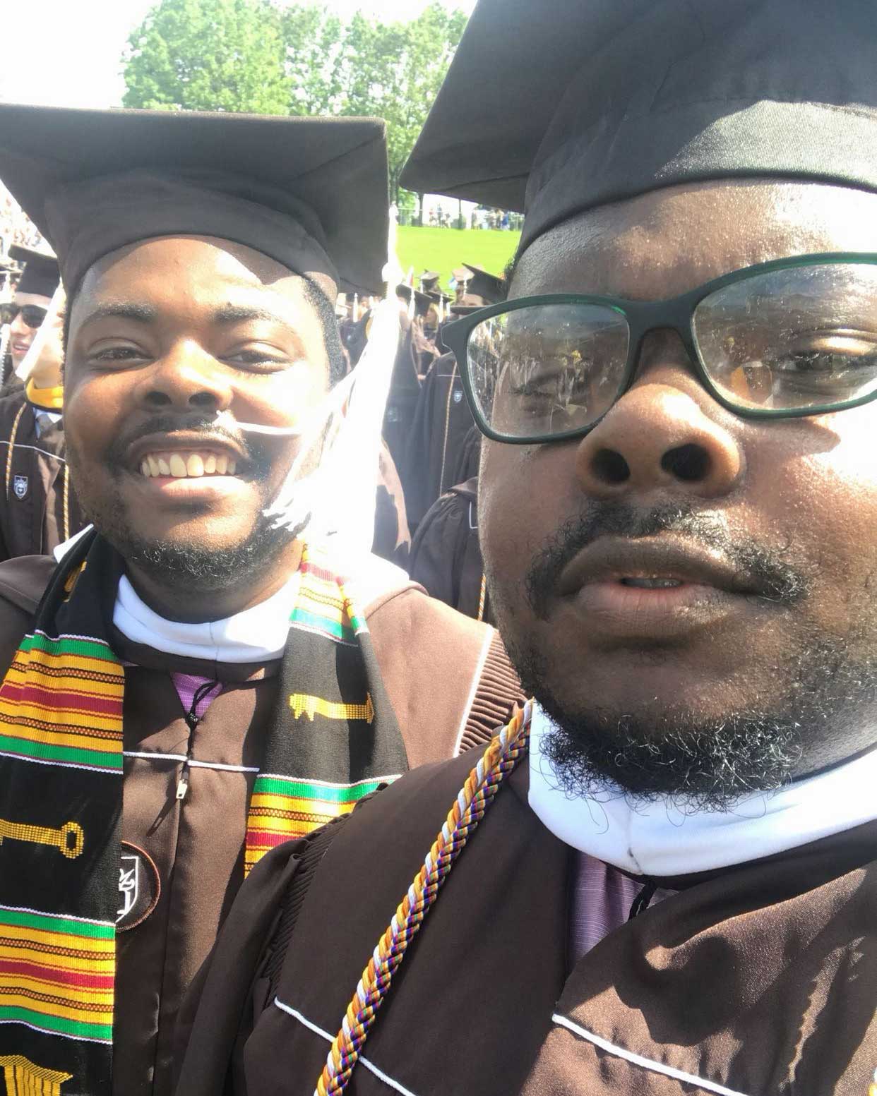 Jamir and Jamal wear brown graduation robes and matching caps. The light is bright and warm; behind them are other students.