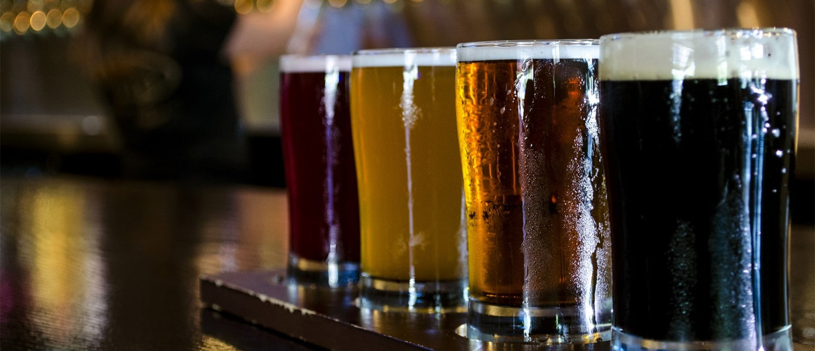 Four perspiring glasses of varying shades of beer with foam on a wooden countertop.