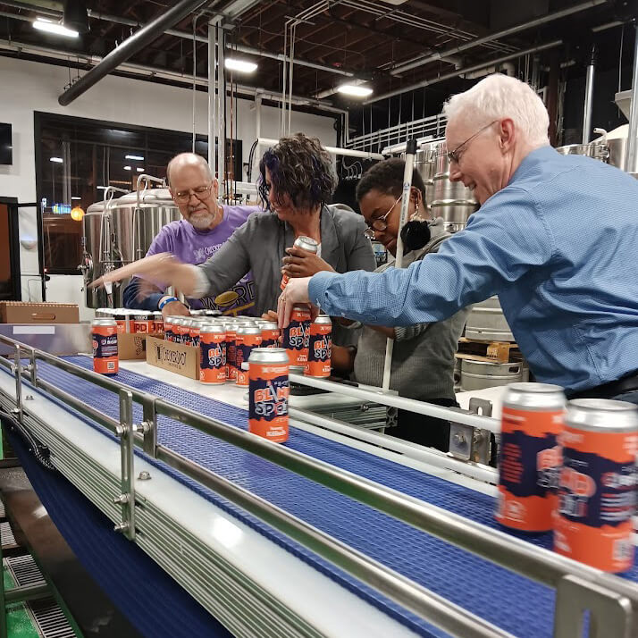 Standing in a production facility with three other BISM associates, Mike Gosse helps move orange label beer cans to a conveyor belt in front of them.