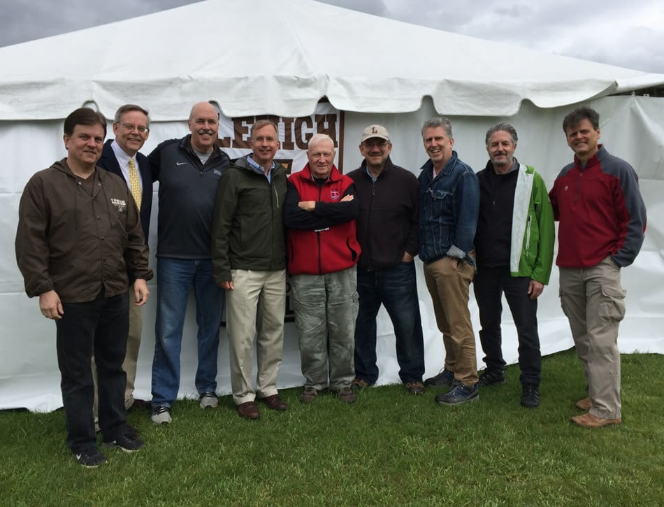 Nine men wearing khakis, jeans, jackets, fleece vests, and pullovers, stand side-by-side outside a white tent with a Lehigh banner hanging on it.