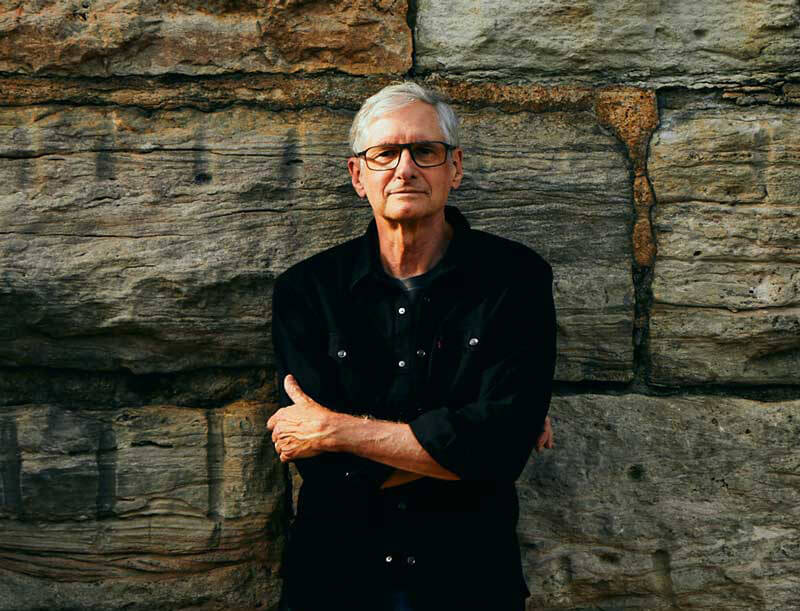 Jim Stanard wears a casual black button down shirt over a t-shirt while crossing his arms and leaning against a large stone wall.