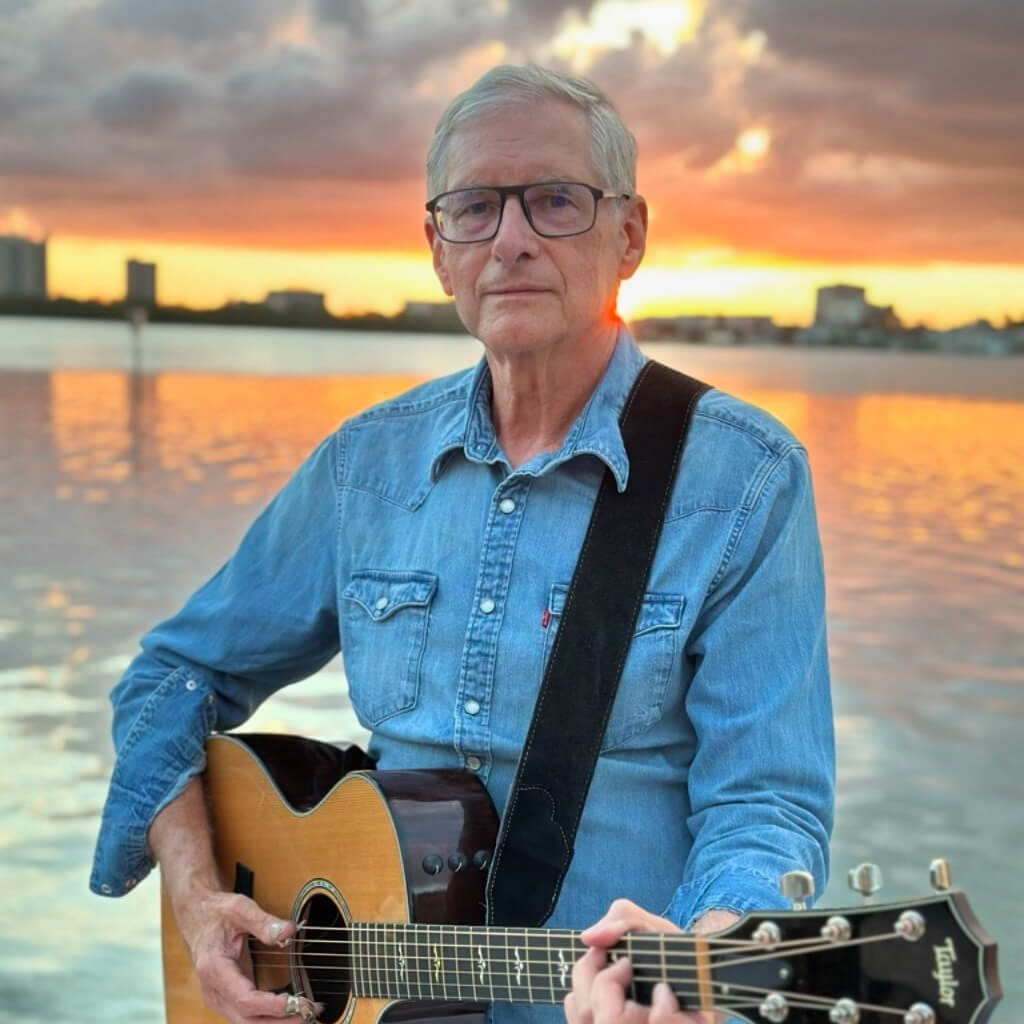 Jim Stanard poses holding a guitar on a dock with the sunset blazing over the water in the distance.