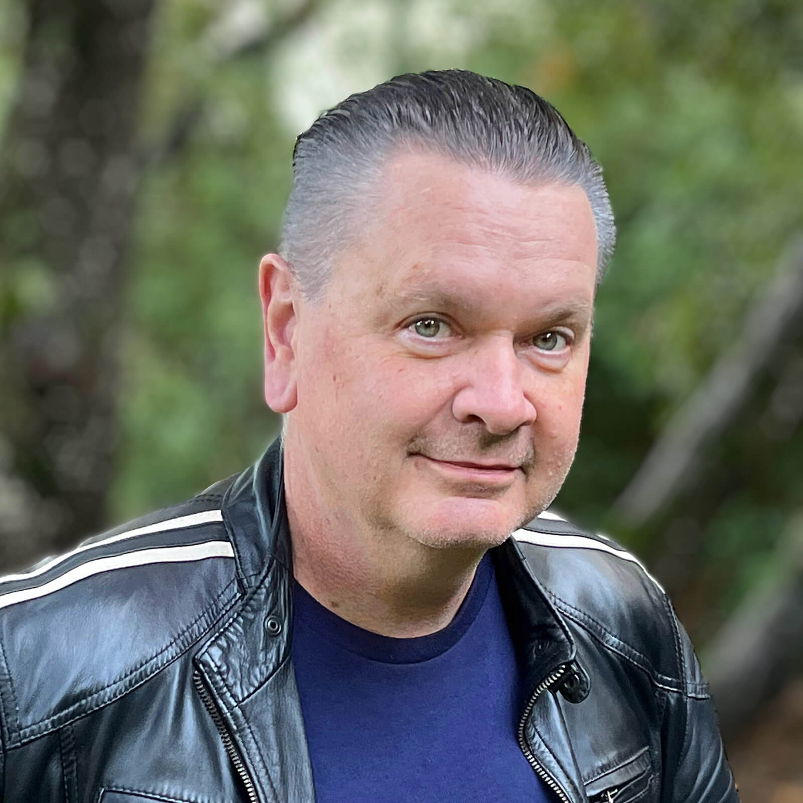 Joe Paluska wears a black leather jacket over a blue shirt and poses with a close mouth smile for a headshot.