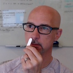 Lippiner holds a Nampon up to his nose. He wears black glasses and behind him is a whiteboard with writing on it.