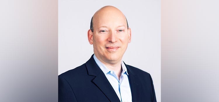 Joshua wears a light blue button up and navy suit jacket; he smiles at the camera
