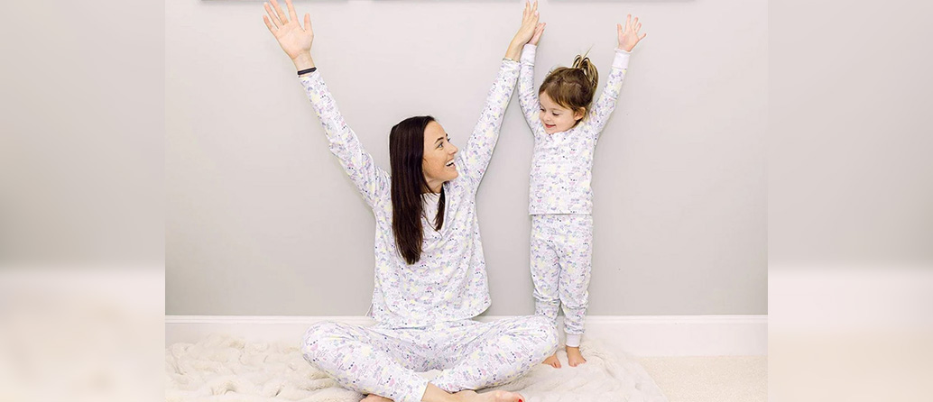 A mother and daughter wearing matching white pajamas smiling at each other with their hands in the air