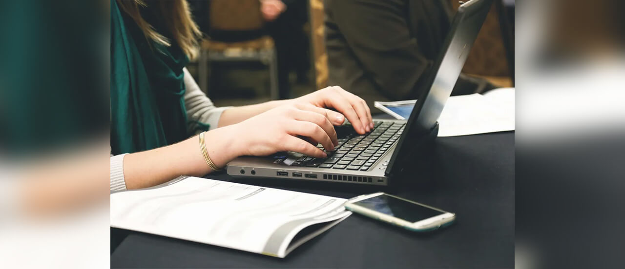 A close up of a person’s hands typing on a laptop with a piece of paper next to it.