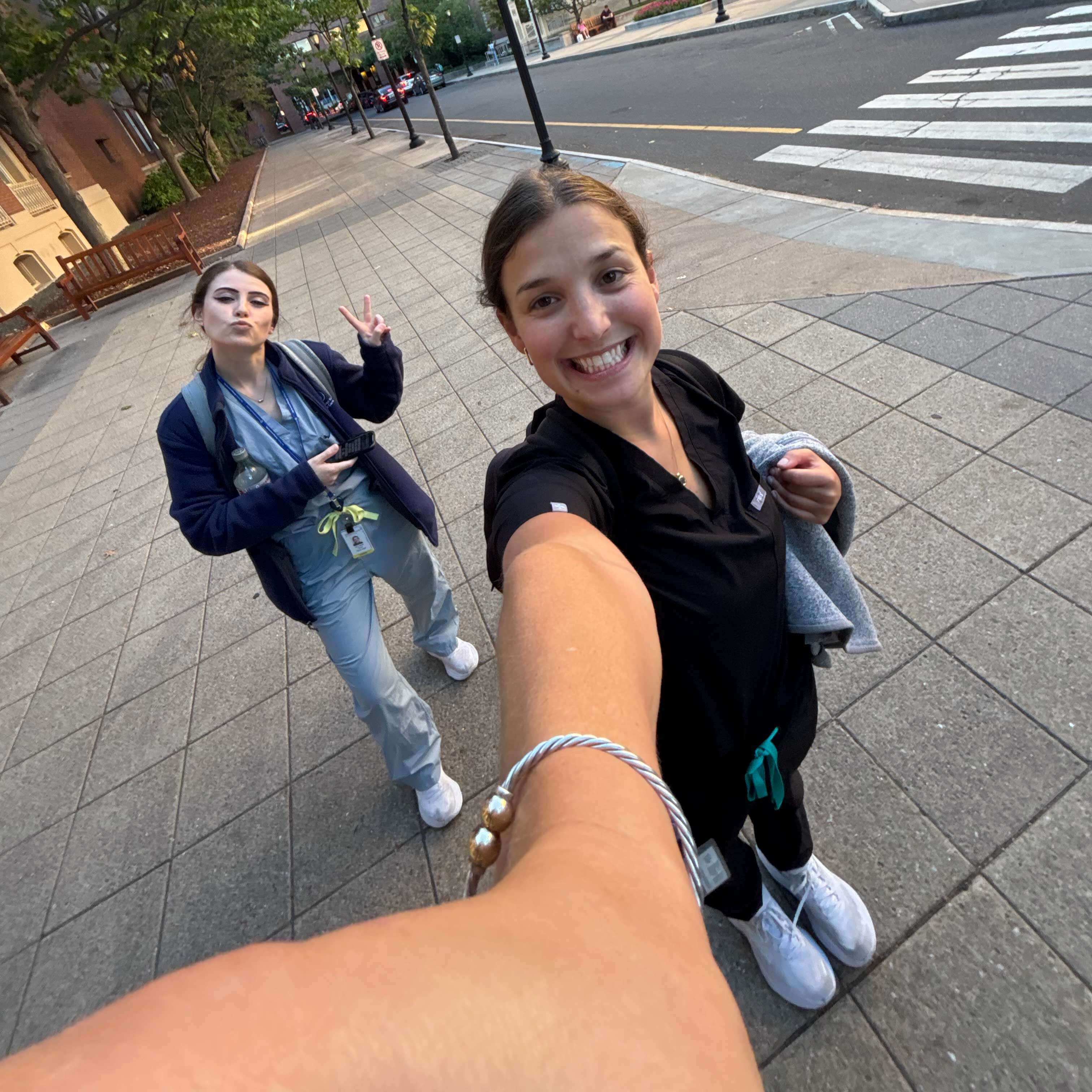 Ali and Julia smile while posing in their scrubs in the city. Ali makes a peace sign with her hand; their scrubs are blue.